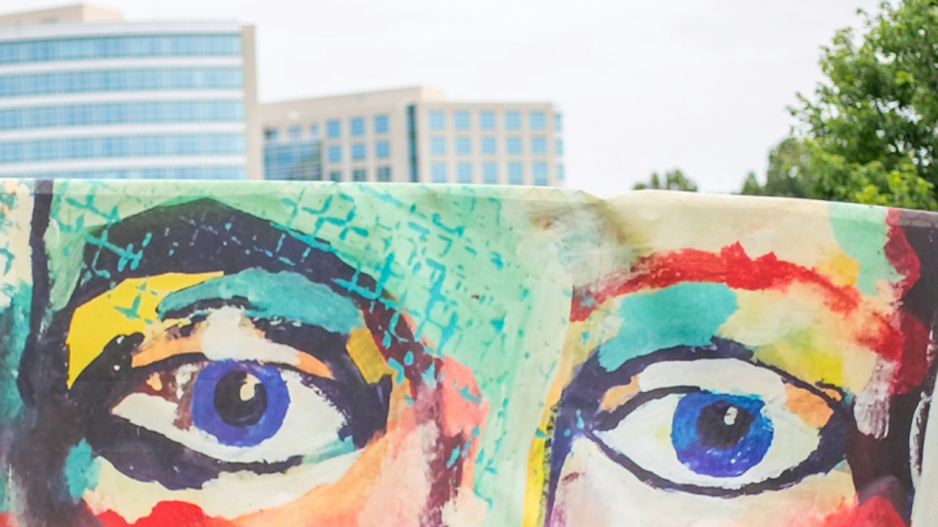 Printed mural on construction fencing depicts a colorful face with large eyes.
